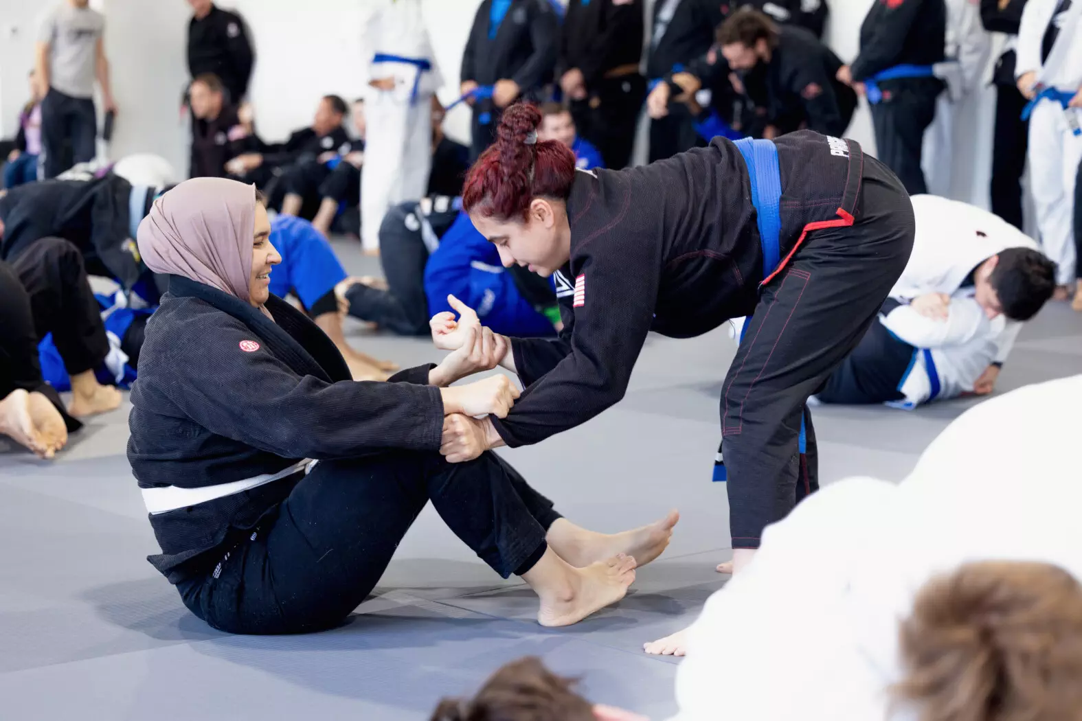 Brazilian Jiu-Jitsu for Women Women training in Brazilian Jiu-Jitsu, one sitting on the ground, the other leaning forward with hands gripping the other's pants